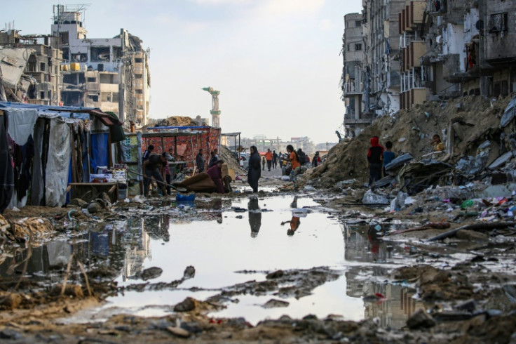 Displaced Palestinians walk around a puddle in front of destroyed buildings and tents in Khan Yunis