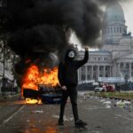 A demonstrator wearing a Guy Fawkes mask walks in front of a Cadena 3 radio station car set on fire during a protest outside the National Congress in Buenos Aires