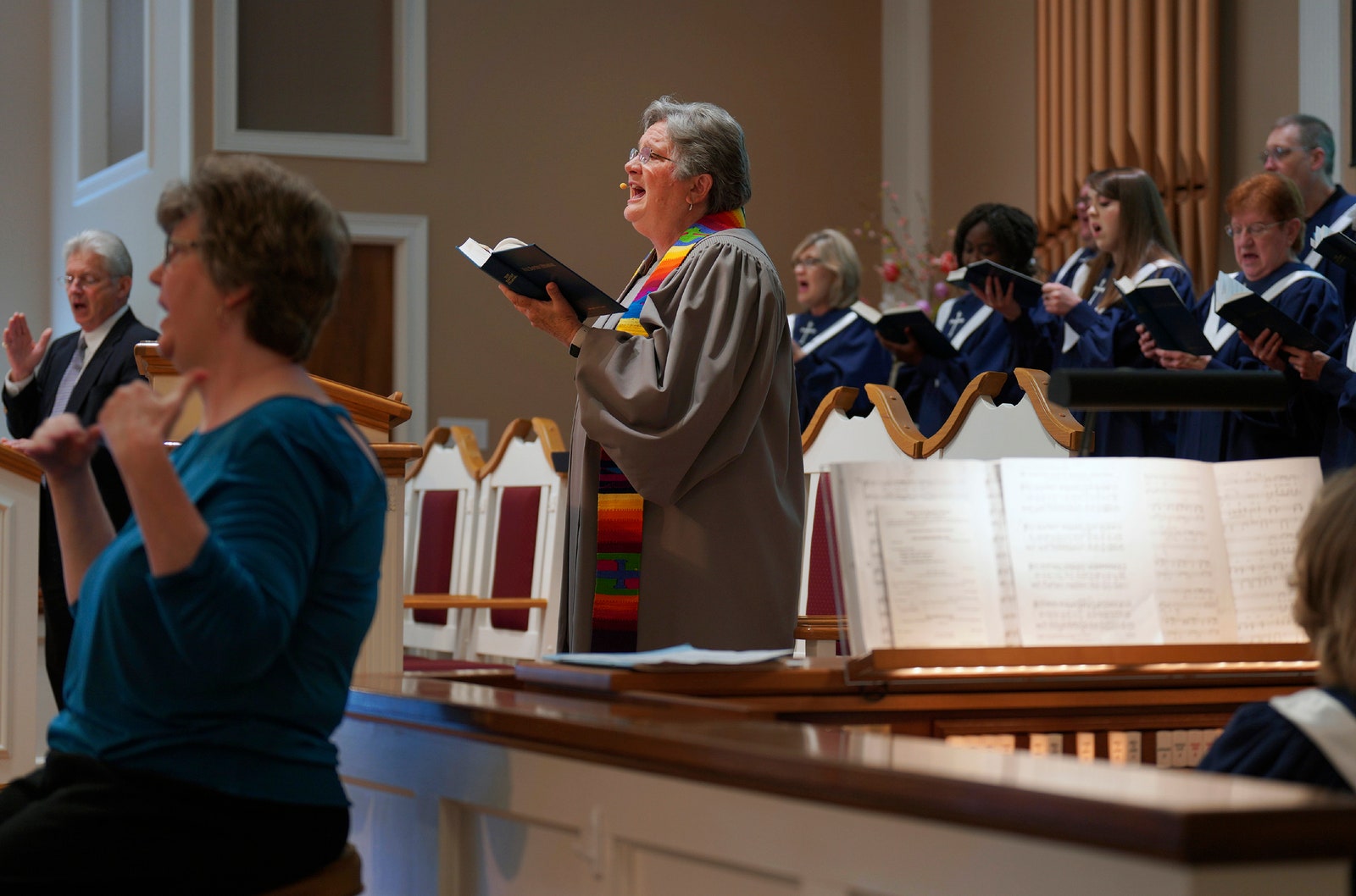 Reverend Linda Barnes Popham standing in front of a choir singing holding a hymnal.