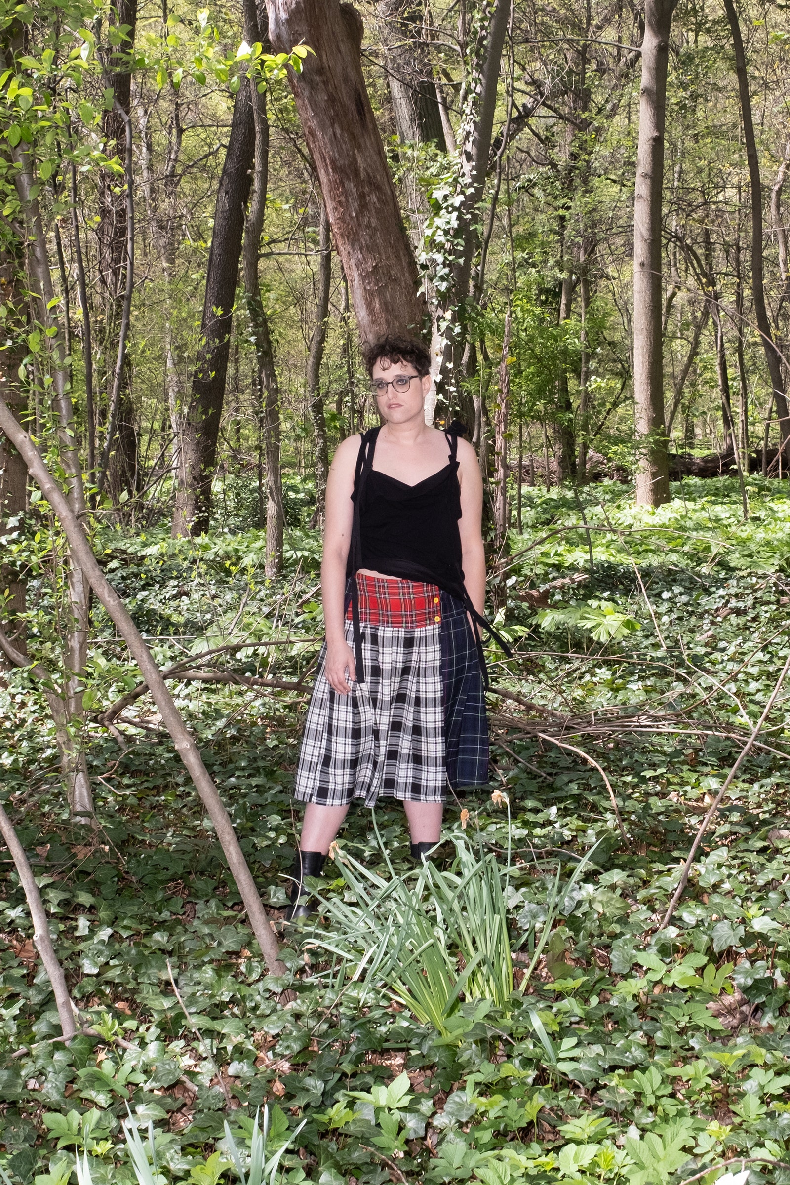 Jane Schoenbrun standing in a wooded area in Prospect Park Brooklyn. They wear a plaid skirt and black tank top.