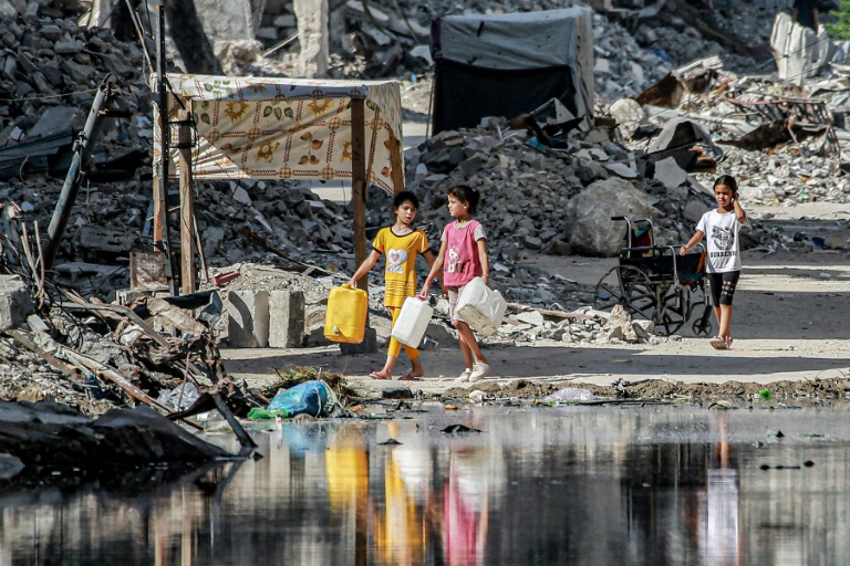 Girls walk with water containers past destroyed buildings and open sewage in Khan Yunis in the southern Gaza Strip on July 8, 2024
