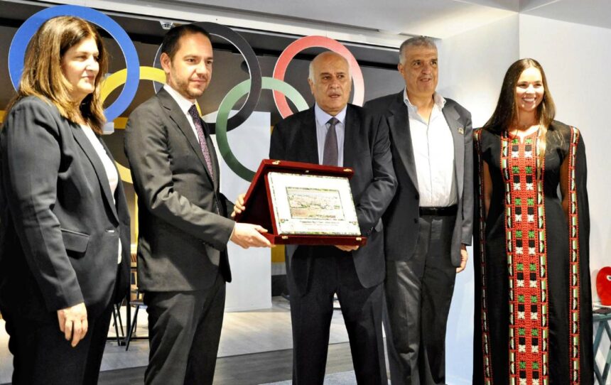 Five people (four in suits, one woman in traditional Palestinian dress) stand behind Olympic rings. The middle two hold a certificate.