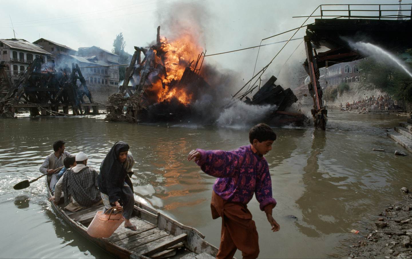 Several men, women, and children use a boat to cross the Jhelum River as the Ali Kadal bridge burns nearby, Srinagar, India, September 1, 1990. The bridge was set on fire by Kashmiri separatists