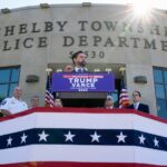 Sen. JD Vance at a podium above bunting, in front of the Shelby Township Police Department, flanked by police officers.